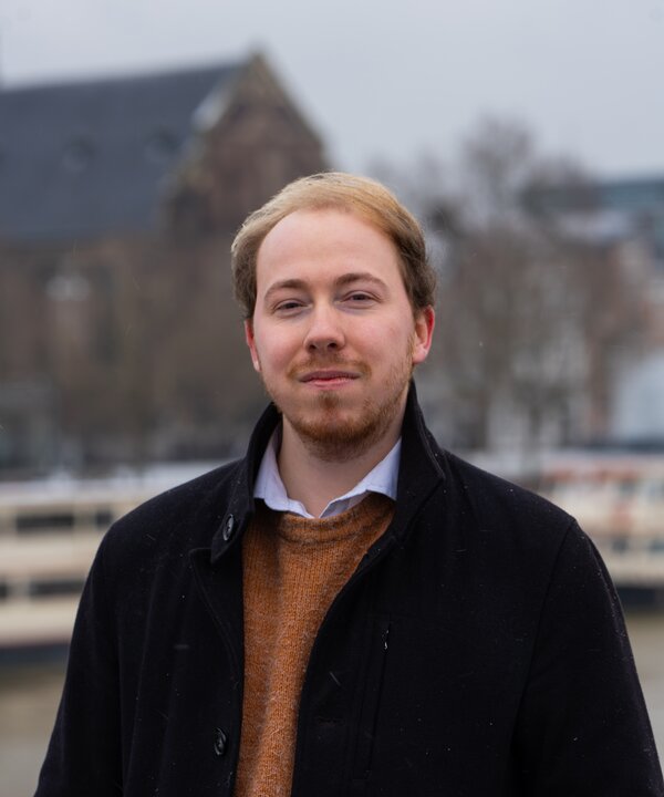 foto van een man met rossig haar, een zwarte jas en bruine trui eronder 